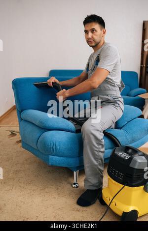 Vertikales Porträt eines professionellen Reinigungsdienstes in voller Länge, der eine Uniform trägt, mit einem Staubsauger, um das blaue Sofa im Wohnzimmer zu reinigen Stockfoto