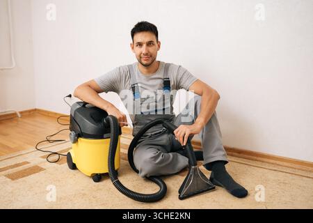 Porträt eines professionellen Reinigungsdienstmitarbeiters, der sich auf dem Boden neben dem Staubsauger befindet und für einen gründlichen Wohnboden bereit ist Stockfoto