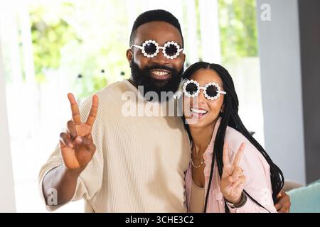 Lächelndes glückliches Paar, das drinnen eine lustige Sonnenbrille trägt, zusammen Friedenszeichen macht, zu Hause Stockfoto