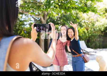 Verschiedene Freundinnen posieren für Smartphone-Fotos im Garten, lächeln und machen Friedenszeichen Stockfoto
