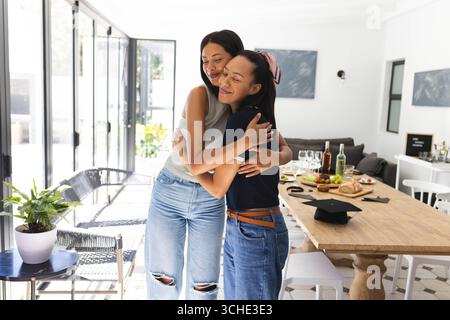 Zwei verschiedene weibliche Freunde umarmen sich zuhause freudig und feiern mit Essen und Getränken auf dem Tisch Stockfoto