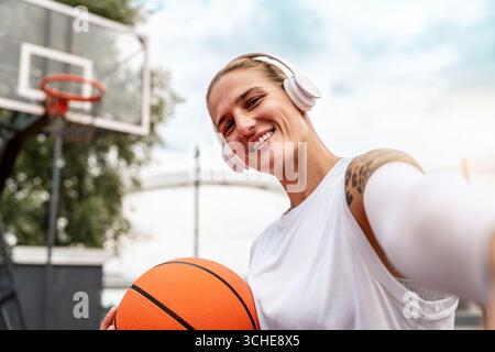 Junge Frau in der Stadt mit schnurlosen Kopfhörern, die Selfie mit Basketball auf dem Straßenplatz macht Stockfoto