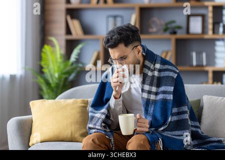 Junger Mann, der auf einem Sofa sitzt, das mit einer Decke bedeckt ist, mit heißem Getränk und Taschentuch. Er scheint krank zu sein, die Nase bläst, in warme Decke gehüllt, und versucht, sich b zu fühlen Stockfoto