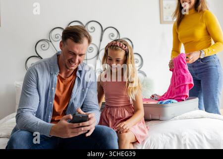 Vater und Tochter lächeln auf das Smartphone, während Mutter den Koffer ins Bett packt Stockfoto