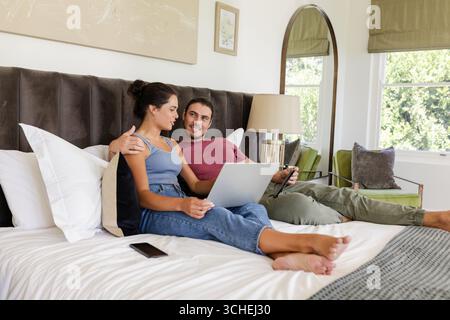 In einem modernen Schlafzimmer mit Messinglampe können Sie ein silbernes Laptop und Smartphone auf dem Bett mit Kopfkissen genießen Stockfoto