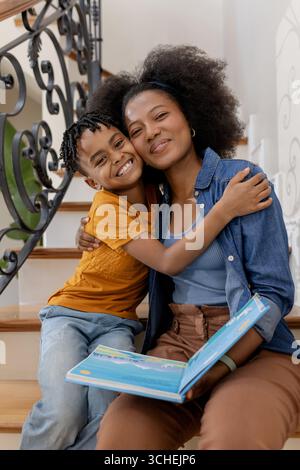 Mutter und Sohn sitzen auf der Treppe, umarmen und lächeln mit offenem Buch Stockfoto