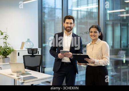 Zwei lächelnde Geschäftsleute halten ein digitales Tablet und Dokumente in der Hand, stehen in einem modernen Büro mit großen Fenstern und zeigen Teamarbeit und Professionalität Stockfoto