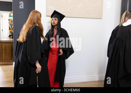 Absolventen in Mützen und Kleidern unterhalten sich nach der Zeremonie aufgeregt zu Hause Stockfoto