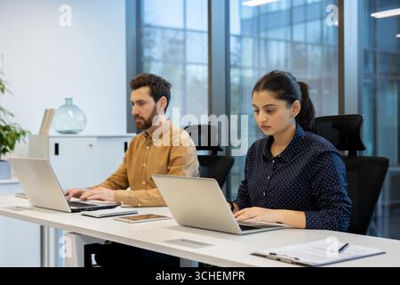 Zwei konzentrierte Büromitarbeiter nutzen Laptops an einem gemeinsamen Schreibtisch in einem hellen, modernen Büro, was Teamarbeit und Produktivität in einer professionellen Umgebung unterstreicht Stockfoto