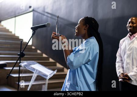 Frau in blauem Hemd, die auf dem Podium spricht, den Preis hält, selbstbewusst lächelt, im Konferenzsaal Stockfoto