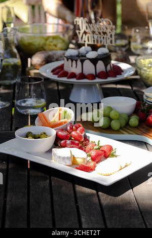 Feierlichkeiten im Freien mit Käseplatte, frischem Obst und Kuchen auf Holztisch Stockfoto