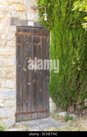 Verwitterte Holztür mit rostigen Scharnieren und grüner Hecke in einem mittelalterlichen Dorf in Drome, Frankreich Stockfoto