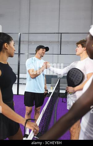 Verschiedene Padel-Spieler schütteln sich die Hände auf einem violetten Platz in der Sporthalle mit Schlägern per Netz Stockfoto