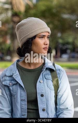 Asiatische Frau, die auf dem Bürgersteig der Stadt steht und rechts mit Beanie, Jeansjacke, Umhängetasche blickt Stockfoto