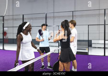 Verschiedene Freunde stehen im Padel Court Netz, plaudern nach dem Spiel mit Paddeln, Feuerschlauchrolle Stockfoto