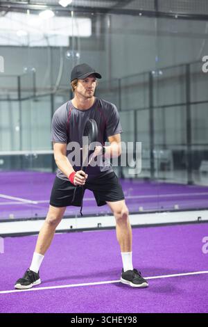 Männlicher Paddeltennisspieler mit Schläger und auf einem violetten Platz neben Glaswänden Stockfoto