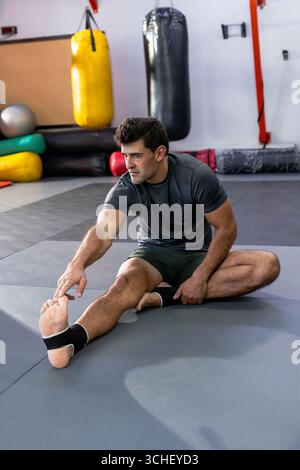 Mann, der die Oberschenkel auf der Kampfkunstmatte im Studio dehnt, mit hängenden Boxsäcken, gestapelten Matten Stockfoto