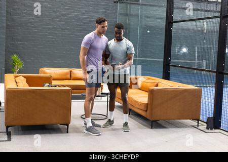 Verschiedene männliche Freunde in sportlicher Kleidung halten blaue Wasserflasche, scrollendes Smartphone in der Lounge Stockfoto