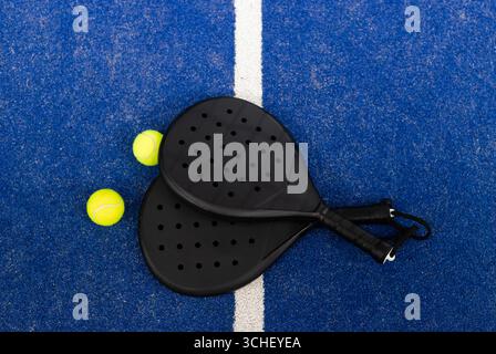 Zwei schwarze Padel-Schläger überlappen sich in der Nähe der weißen Feldlinie auf dem blauen Platz mit Tennisbällen Stockfoto