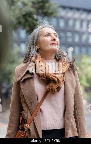 Ältere Frau in braunem Mantel und braunem Strickschal mit einer Umhängetasche aus Leder auf dem Urban plaza Stockfoto