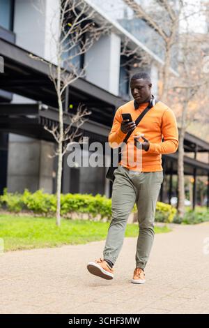 afroamerikanischer Mann, der über einen gepflasterten Weg mit Kopfhörern läuft, indem er ein Smartphone benutzt, das Kaffee hält Stockfoto