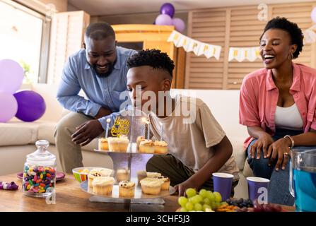 Auf einer Party, bei der er zu Hause Geburtstag feiert, bläst ein afroamerikanischer Junge Kerzen auf Cupcakes aus Stockfoto