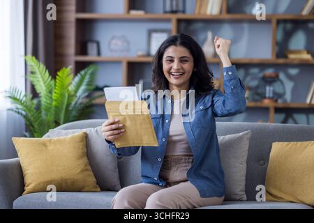Porträt einer jungen Indianerin, die zu Hause auf dem Sofa sitzt. Einen offenen Umschlag mit einem Brief in der Hand halten und sich über den Erfolg freuen und sie großziehen Stockfoto