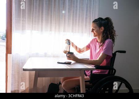 Glückliche Frau mit Behinderung, die Kaffee in ihre Tasse gießt, an einem Tisch in ihrem Haus sitzt Stockfoto