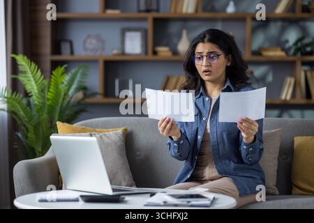 Besorgte junge Indianerin sitzt zu Hause auf der Couch und sieht schockiert auf die Dokumente aus, die sie hält. Stockfoto