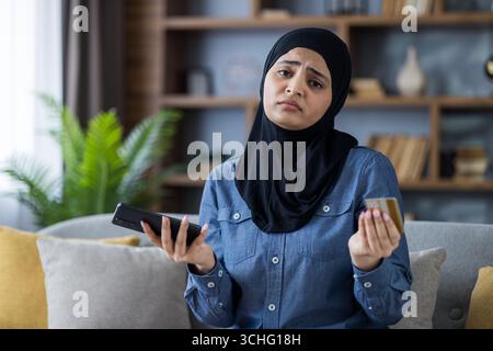 Porträt einer besorgten und enttäuschten jungen Frau im Hijab, die zu Hause auf dem Sofa sitzt, Kreditkarte und Telefon hält und vor der Kamera die Arme spreizt. Stockfoto