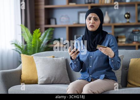 Verärgert und enttäuscht junge muslimische Frau im Hijab, die zu Hause auf dem Sofa sitzt, Kreditkarte und Telefon hält und in die Kamera schaut. Stockfoto
