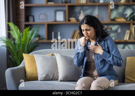 Die junge indische Frau sitzt zu Hause auf der Couch und hustet, bedeckt ihren Mund mit der Hand und hält ihre Brust. Stockfoto