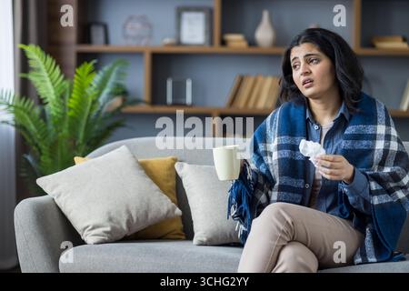 Müde und kranke junge indische Frau, die zu Hause auf der Couch sitzt, in eine Decke gewickelt, einen Becher mit einem Getränk und einer Serviette hält und an dem Virus leidet Stockfoto