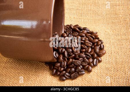 Nahaufnahme von dunkel gerösteten Kaffeebohnen, die aus einer braunen Keramikschale auf einen rustikalen Sacksack fließen und einen warmen, aromatischen und strukturierten Hintergrund schaffen Stockfoto
