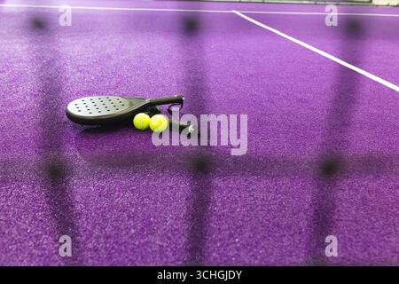 Schwarzer Padel-Schläger, der neben zwei Tennisbällen auf einem violetten Platz hinter verschwommenem Netz liegt Stockfoto