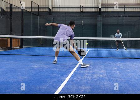 Verschiedene Männer, die sich auf dem Padel-Platz versammeln, schlagen mit einem Paddel einen kalkgrünen Ball neben dem Gitterzaun Stockfoto