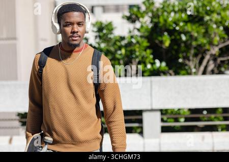 afroamerikanischer Mann, der durch die Stadt geht, Skateboard trägt, Rucksack trägt Stockfoto