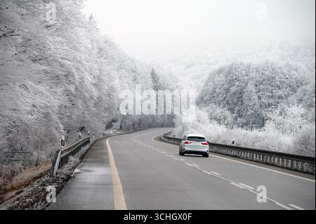 Ein Auto fährt auf einer schneebedeckten Straße durch eine ruhige weiße Winterlandschaft, umgeben von satinierten Bäumen und Feldern in der Weihnachtszeit Stockfoto