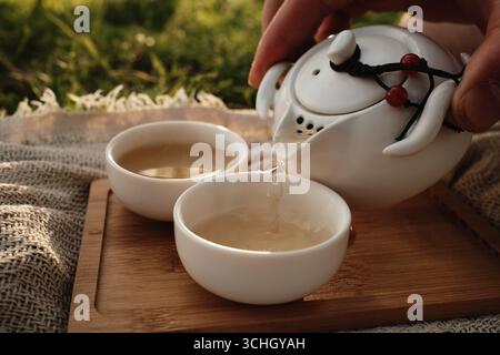 Geben Sie heissen Tee von Hand aus einer weißen Teekanne in eine kleine Porzellantasse draußen auf einem Holztablett. Picknick im chinesischen Stil für zwei Personen im Freien. Stockfoto
