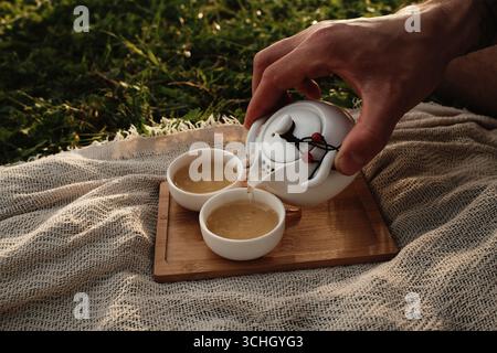 Geben Sie heissen Tee von Hand aus einer weißen Teekanne in eine kleine Porzellantasse draußen auf einem Holztablett. Picknick im chinesischen Stil für zwei Personen im Freien. Stockfoto