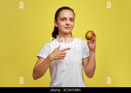 Glückliche Frau in weißem T-Shirt, die Kiwi auf gelbem Studiohintergrund hält Stockfoto