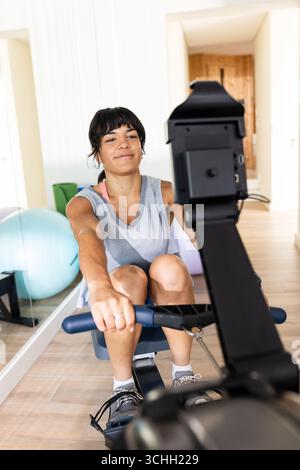 Frau, die zu Hause auf dem Rudergerät trainiert, lächelt und genießt das Training Stockfoto