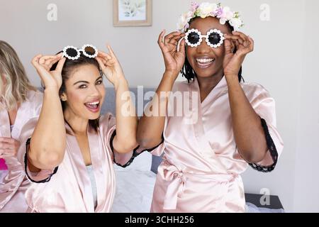 Verschiedene Freundinnen in rosafarbenen Bademänteln, die zu Hause eine Sonnenbrille anprobieren und fröhlich lächeln Stockfoto