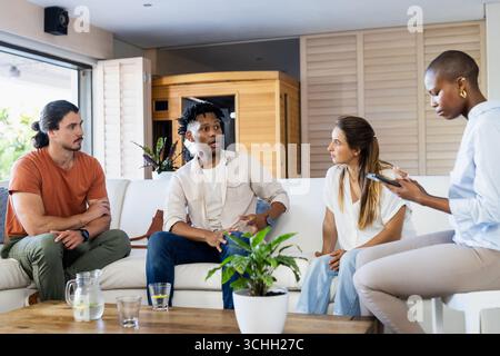 Verschiedene Freunde diskutieren zu Hause, einer nutzt das Smartphone, andere hören aufmerksam zu Stockfoto
