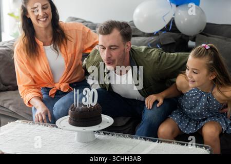 Er feiert Geburtstag und bläst Kerzen auf Kuchen mit Tochter und Partner aus Stockfoto