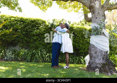 Bei der Hochzeit, ein älteres Paar, das sich unter einem Baum im Garten umschließt und den sonnigen Tag zusammen genießt Stockfoto