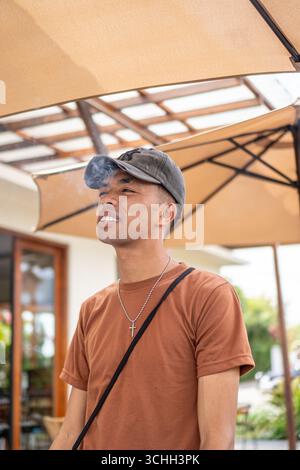 Ein ungezwungener junger Mann in braunem T-Shirt und Mütze raucht draußen auf einer Café-Terrasse. Stockfoto