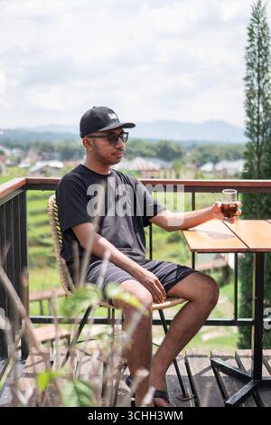 Ein asiatischer Mann mit schwarzem T-Shirt und Mütze sitzt auf einer Terrasse und trinkt Kaffee Stockfoto