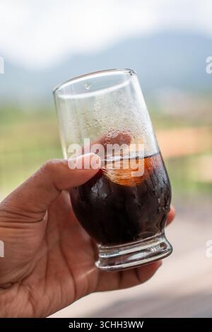 Nahaufnahme einer Hand, die ein Glas eiskalten schwarzen Kaffees mit sichtbaren Eiswürfeln und Kondensation auf dem Glas hält. Stockfoto