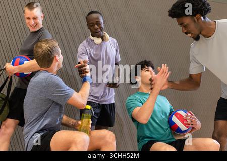 Verschiedene männliche Volleyballpartner ruhen in der Sporthalle aus und halten Volleybälle und eine Wasserflasche Stockfoto